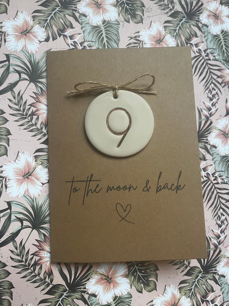 May include: A brown card with the text "to the moon & back" and a heart symbol. A small, round, beige ceramic ornament with the number 9 is attached to the card with twine.