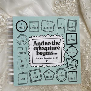 May include: A light blue spiral-bound anniversary book with white wire binding. The cover features the text 'And so the adventure begins... The Anniversary Book' surrounded by sixteen numbered boxes listing traditional anniversary materials from paper to wax.