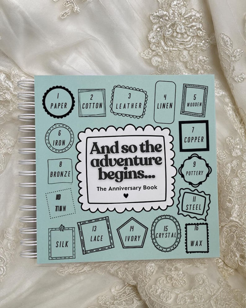 May include: A light blue spiral-bound anniversary book with white wire binding. The cover features the text 'And so the adventure begins... The Anniversary Book' surrounded by sixteen numbered boxes listing traditional anniversary materials from paper to wax.
