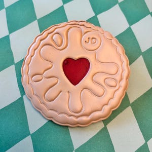 May include: A round, tan-coloured biscuit with a red heart-shaped centre. The biscuit has a textured edge and a design of flowing shapes. The initials "JD" are embossed on the top. The biscuit is on a green and white chequered surface.