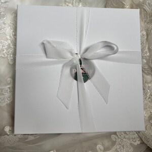 May include: A square white gift box tied with a white ribbon bow. A circular tag with the text "THE PEONY CLUB" is attached to the centre of the ribbon. The box has a smooth matte finish and is centred on a white lace fabric background.