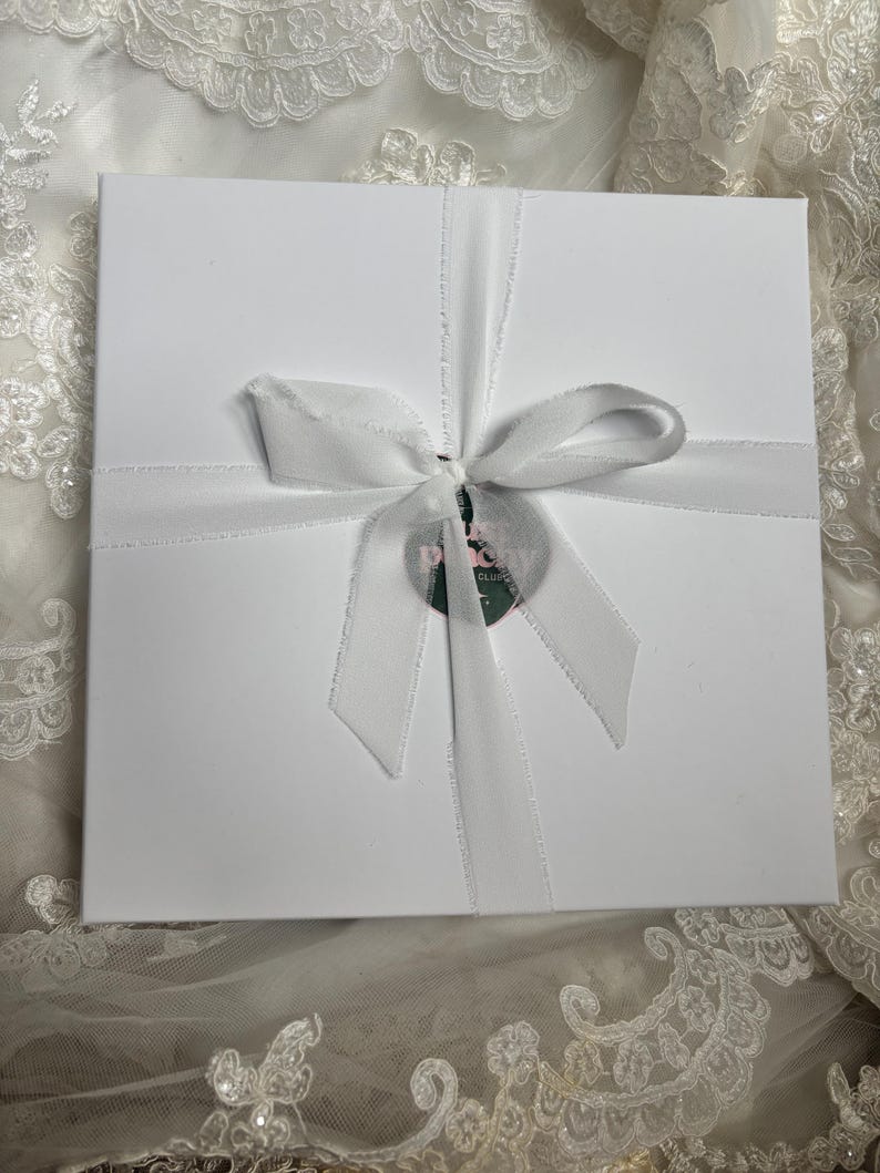 May include: A square white gift box tied with a white ribbon bow. A circular tag with the text "THE PEONY CLUB" is attached to the centre of the ribbon. The box has a smooth matte finish and is centred on a white lace fabric background.