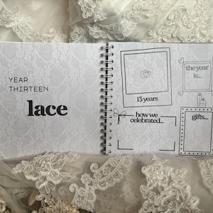 May include: A spiral-bound anniversary scrapbook with a white lace-patterned background. The left page reads YEAR THIRTEEN lace. The right page features hand-drawn frames for photos and text, including labels for 13 years, the year is, how we celebrated, and gifts.