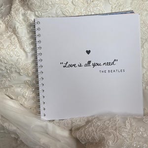 May include: A square white spiral-bound notebook with a black heart icon and the text "Love is all you need" followed by "THE BEATLES" printed on the front cover, resting on top of a white lace fabric with intricate floral embroidery patterns.