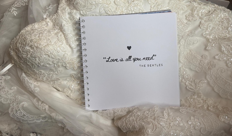 May include: A square white spiral-bound notebook with a black heart icon and the text "Love is all you need" followed by "THE BEATLES" printed on the front cover, resting on top of a white lace fabric with intricate floral embroidery patterns.