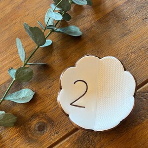 May include: White ceramic table number holder shaped like a cloud with the number "2" written in black. The holder has a textured surface on one side.