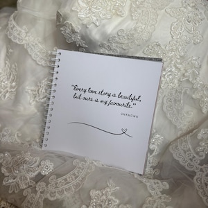 May include: A square white spiral-bound notebook featuring the black printed text, "Every love story is beautiful, but ours is my favourite." followed by the word "UNKNOWN" and a simple line drawing of a heart at the end of a curved line.