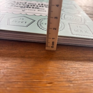 May include: A light mint green anniversary book titled And so the adventure begins with various labels including 14 Ivory, 16 Wax, and Steel. A wooden ruler is placed vertically against the book, showing a thickness of approximately 1.3 centimetres.