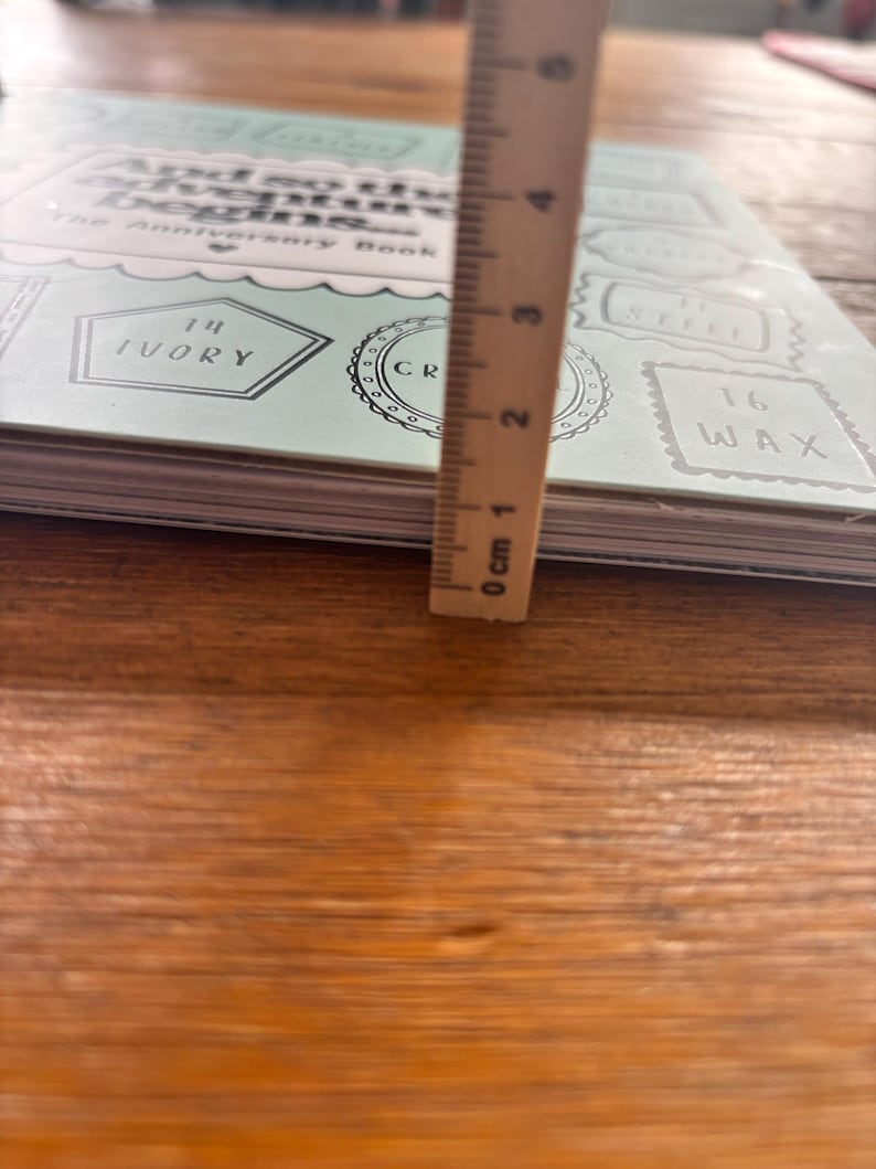 May include: A light mint green anniversary book titled And so the adventure begins with various labels including 14 Ivory, 16 Wax, and Steel. A wooden ruler is placed vertically against the book, showing a thickness of approximately 1.3 centimetres.