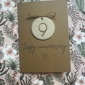 May include: A brown card with the text "happy anniversary" and a heart symbol. A white ceramic ornament with the number 9 is attached to the card with twine.