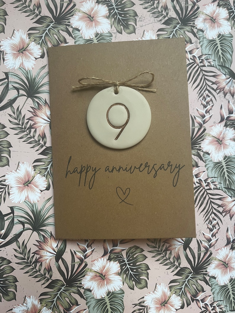 May include: A brown card with the text "happy anniversary" and a heart symbol. A white ceramic ornament with the number 9 is attached to the card with twine.