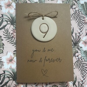 May include: A brown card with a white ceramic ornament attached with twine. The ornament has the number 9 inscribed on it. The card reads "you & me... now & forever" with a heart symbol.