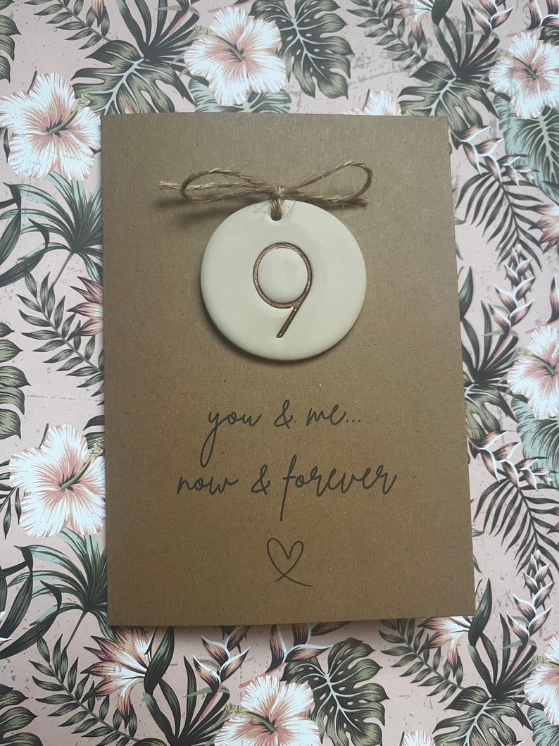 May include: A brown card with a white ceramic ornament attached with twine. The ornament has the number 9 inscribed on it. The card reads "you & me... now & forever" with a heart symbol.
