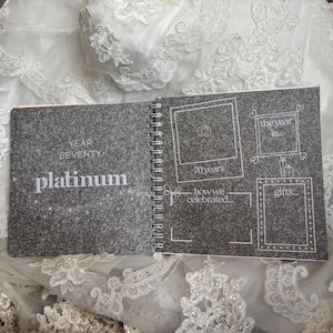 May include: A spiral-bound memory book with a dark grey glitter cover featuring the text YEAR SEVENTY platinum. The interior page has white line-drawn frames and text including 70 years, the year is, how we celebrated, and gifts, all on a dark grey background.
