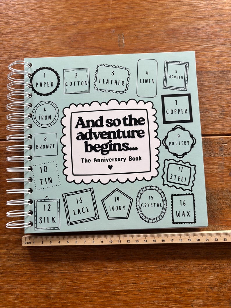 May include: A light blue spiral-bound anniversary book with the text "And so the adventure begins... The Anniversary Book" and sixteen numbered frames listing materials from paper to wax, placed next to a ruler measuring 23 centimetres in length.