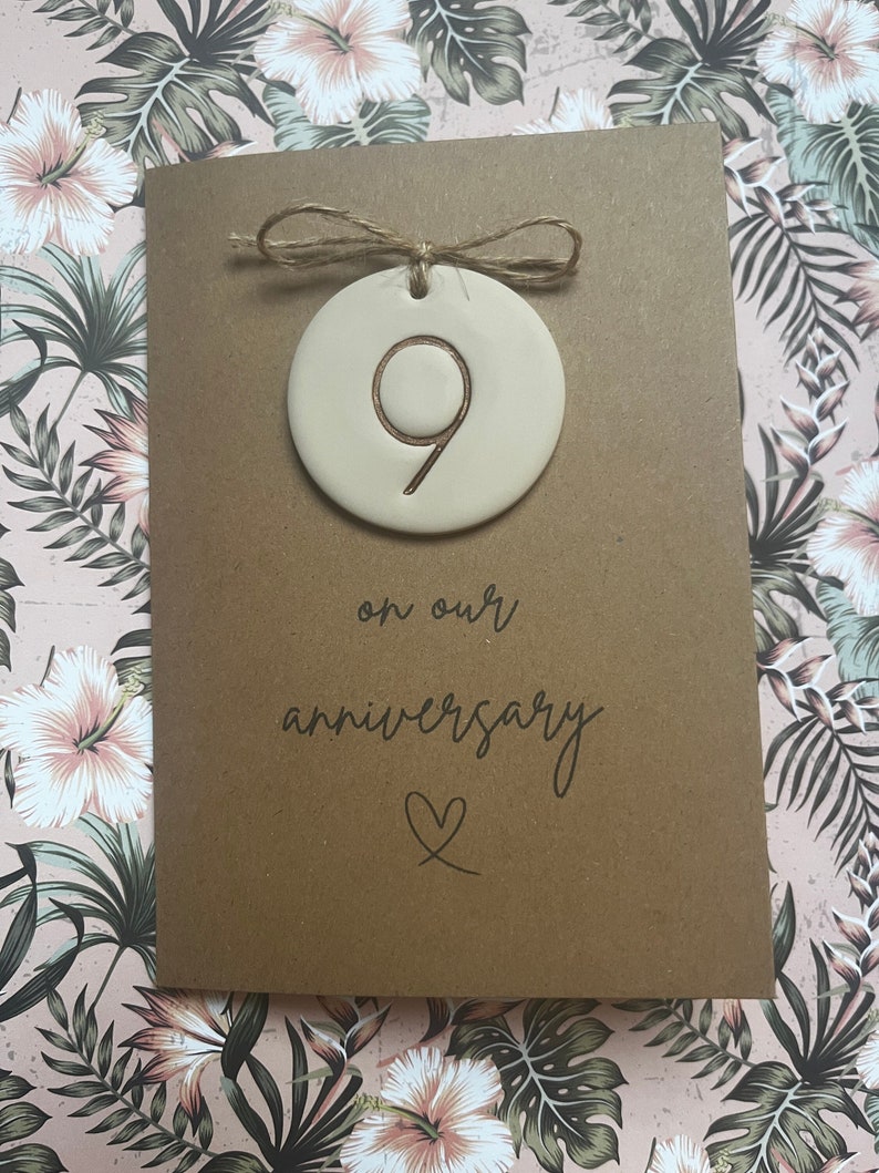 May include: A brown card with the text "on our anniversary" and a heart symbol. A small, round, beige ceramic ornament with the number "9" inscribed on it is attached to the card with twine.