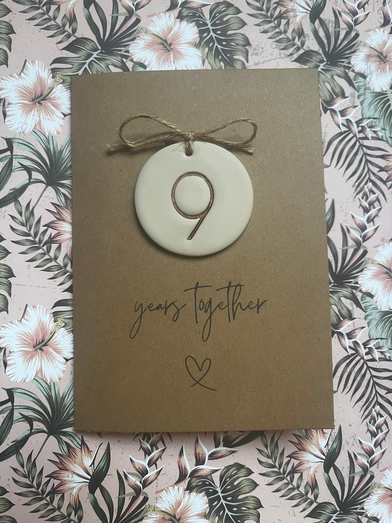 May include: A brown card with the text "9 years together" and a heart symbol. A small, round, beige ceramic ornament with the number "9" inscribed in gold is attached to the card with twine.