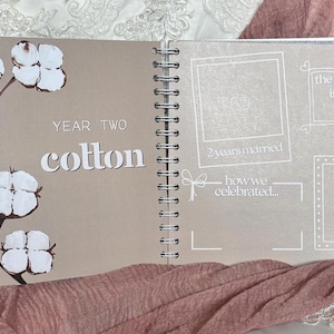 May include: A spiral-bound anniversary scrapbook open to a page titled Year Two Cotton. The left side features a brown background with white cotton boll illustrations. The right side has white line-drawn frames for photos and text including the year is, 2yearsmarried, how we celebrated, and gifts.