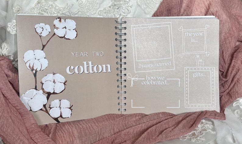 May include: A spiral-bound anniversary scrapbook open to a page titled Year Two Cotton. The left side features a brown background with white cotton boll illustrations. The right side has white line-drawn frames for photos and text including the year is, 2yearsmarried, how we celebrated, and gifts.