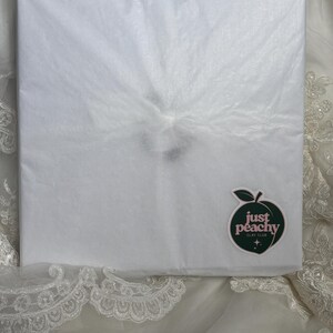 May include: A square package wrapped in white tissue paper. A dark green peach-shaped sticker is affixed to the lower right corner, featuring the text "just peachy CLAY CLUB" in white lettering with two small star icons below the text.
