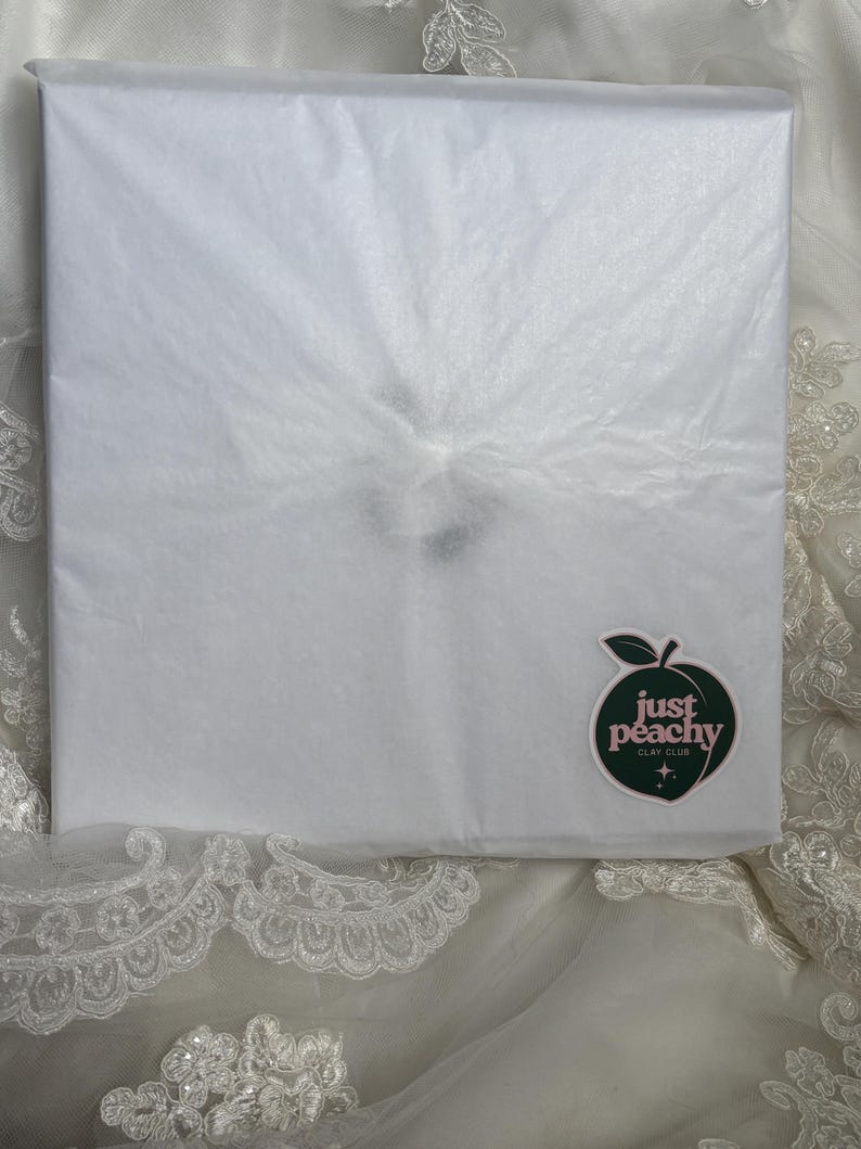 May include: A square package wrapped in white tissue paper. A dark green peach-shaped sticker is affixed to the lower right corner, featuring the text "just peachy CLAY CLUB" in white lettering with two small star icons below the text.