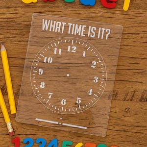 May include: A clear acrylic clock face with the text "WHAT TIME IS IT?" and a clock face outline. Colorful foam letters and numbers are scattered around the clock. Two yellow pencils with pink erasers are also present.