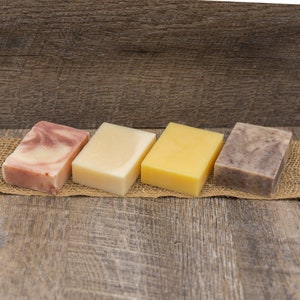 May include: Four bars of soap on a burlap surface. The bars are arranged from left to right: pink and white, white, yellow, and brown. The soap bars are on a wooden background.