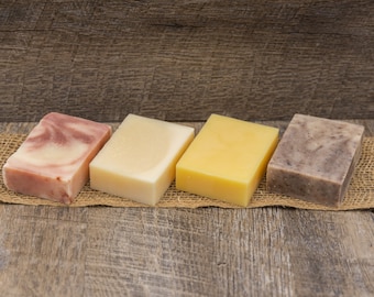 May include: Four bars of soap on a burlap surface. The bars are arranged from left to right: pink and white, white, yellow, and brown. The soap bars are on a wooden background.