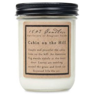May include: A white candle in a glass jar with a black lid. The label reads "1603 Bandlese Apothecary of Fragrant Garde Cabin on the Hill Simple and peaceful the cabin looks on the hill. An American flag stands stately at the forest door. Leaves are swirling around the grass and fresh cut firewood fills the air."