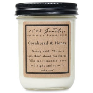 May include: A glass jar candle with a brown label that reads "1803 Candles Apothecary of Fragrant Goods Cornbread & Honey". The label also includes a quote: "Nanny said, "There's somethin' about cornbread, folks eat it mornin' noon and night and some in between.""