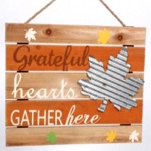 May include: A wooden sign with a rustic, distressed look. It has a brown and orange background with the words "Grateful hearts gather here" in white lettering. A metal leaf with a corrugated texture is attached to the sign.
