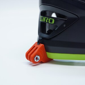 Puede incluir: Primer plano de un casco negro con una franja verde y un soporte GoPro. El soporte es naranja y tiene un tornillo plateado.