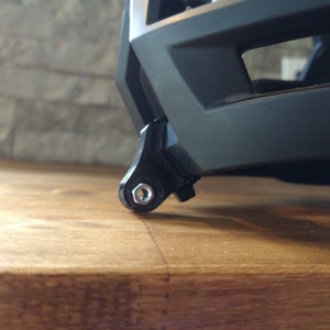 May include: A black helmet with a built-in mount for a camera or action camera. The mount is made of black plastic and has a silver screw.