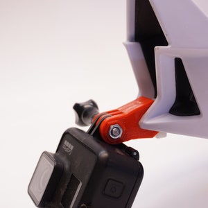 May include: A black GoPro camera mounted on a white helmet with an orange 3D printed mount.
