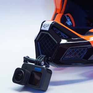 May include: A black and orange motorcycle helmet with a GoPro camera mounted on the front. The helmet has a black visor and a white Fox logo on the front.
