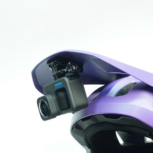 May include: A purple helmet with a visor and a GoPro camera mounted on top. The camera is facing forward and is ready to record.