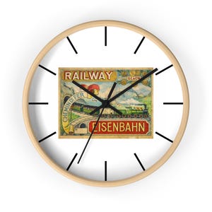 Puede incluir: Un reloj de pared redondo con marco de madera clara y manecillas negras. La esfera del reloj presenta un anuncio de ferrocarril vintage con las palabras "RAILWAY" y "EISENBAHN". El reloj tiene marcadores de hora negros.