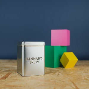 May include: A silver metal tin with the text "HANNAH'S BREW" printed on the front. The tin is sitting on a wooden surface in front of a stack of colorful blocks.