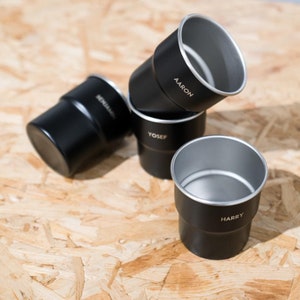 Puede incluir: Cuatro tazas de metal negro con bordes plateados apiladas sobre una superficie de madera. Las tazas están etiquetadas con los nombres "Aaron", "Yosef", "Harry" y "Benjamin".