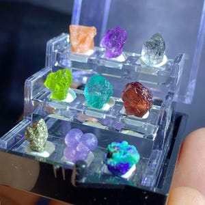 以下が含まれることがあります： 9つのカラフルな原石が入った小さな透明なディスプレイケース。原石はオレンジ、紫、緑、青、茶色など様々な色があります。ケースは黒と透明で、石を展示するための複数の棚があります。
