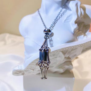 May include: Silver-toned necklace featuring a pendant shaped like a skeletal hand. The necklace has a delicate chain and a small, round bead at the top of the pendant. The pendant's fingers are articulated, giving it a realistic appearance. The necklace is displayed on a white bust.