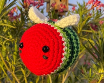 Peut inclure: Ornement fait à la main en crochet représentant une abeille, avec une tête rouge, un corps rayé vert et blanc, et des ailes blanches. L'abeille a des yeux noirs et est suspendue devant un nichoir en bois et du feuillage vert.