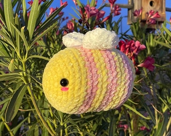 Peut inclure: Un ornement d'abeille fait à la main, avec des rayures jaunes et roses, des yeux noirs et une bouche rose. L'abeille est suspendue devant un feuillage vert, des fleurs roses et un nichoir en bois sur fond de ciel bleu.