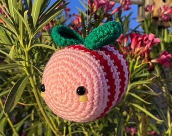 Peut inclure: Un ornement d'abeille crocheté à rayures roses et rouges, avec des ailes vertes et des yeux noirs. L'abeille est suspendue devant un fond de feuillage vert, de fleurs roses et d'un nichoir en bois sur fond de ciel bleu.