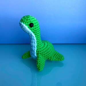 May include: A handmade green crochet Nessie monster with a light blue neck and black safety eyes. The amigurumi Nessie has a long neck, small flippers, and a rounded body. The toy is a cute, whimsical representation of the Loch Ness Monster.