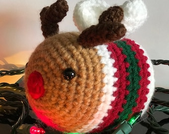Peut inclure: Ornement de Noël fait à la main en crochet représentant un renne avec des détails marron, blancs et rouges. Le renne a un nez rouge, des bois bruns et des yeux noirs. L'ornement est orné de rayures vertes, rouges et blanches et entouré de guirlandes lumineuses.