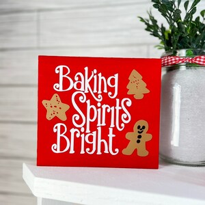 May include: A red wooden sign with the words "Baking Spirits Bright" in white script. The sign features gingerbread man, star, and Christmas tree cutouts. A jar of white powder and greenery are in the background.