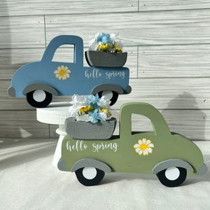 May include: Two wooden truck figurines with a flower arrangement in the bed. The trucks are painted in a light blue and a green color. The trucks have the words "hello spring" written on them.