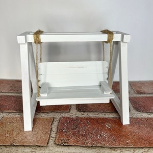 May include: A miniature white wooden swing set with a rope seat. The swing set is on a brick surface.