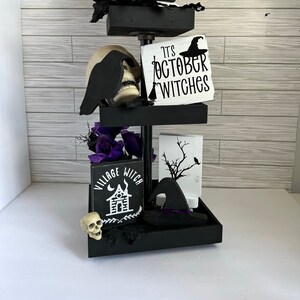 May include: A three-tiered black tiered tray decorated for Halloween. The top tier has a stack of three white and black wooden signs that read "Spells", "Potions", and "Magic". The middle tier has a black raven figurine, a white skull, and a white wooden sign that reads "It's October Witches". The bottom tier has a black witch's hat, a white wooden sign that reads "Village Witch", a white skull, and purple flowers.